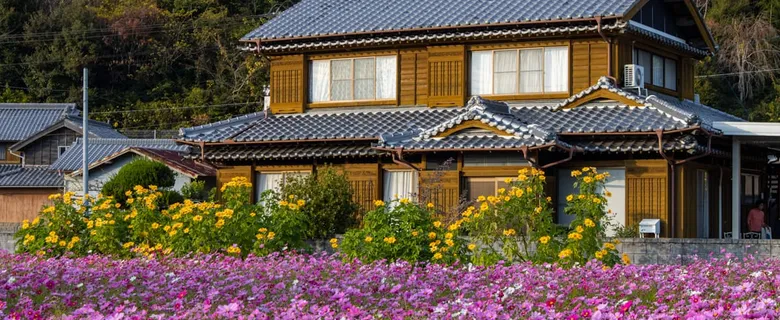 コスモス畑の奥に建つ瓦屋根の木造二階建て住宅
